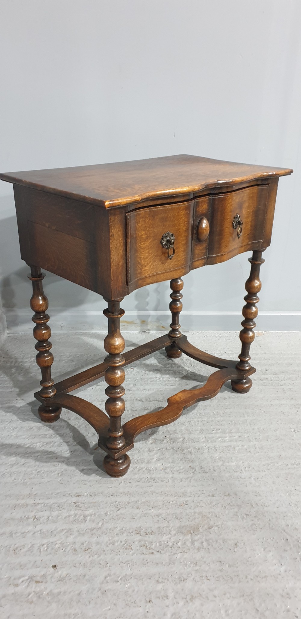 An antique oak side table featuring bobbin turned legs, a shaped frieze drawer with brass drop handles, and a wavy cross-stretcher base. View VI
