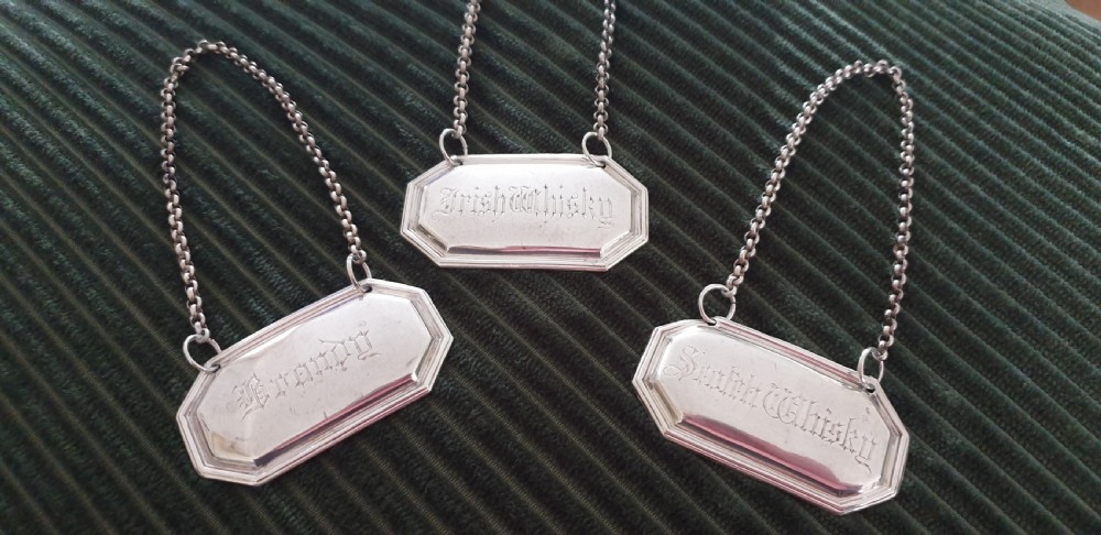 A set of three octagonal silver decanter labels with fine link chains, engraved with Brandy, Irish Whisky, and Scotch Whisky in Gothic script. View V
