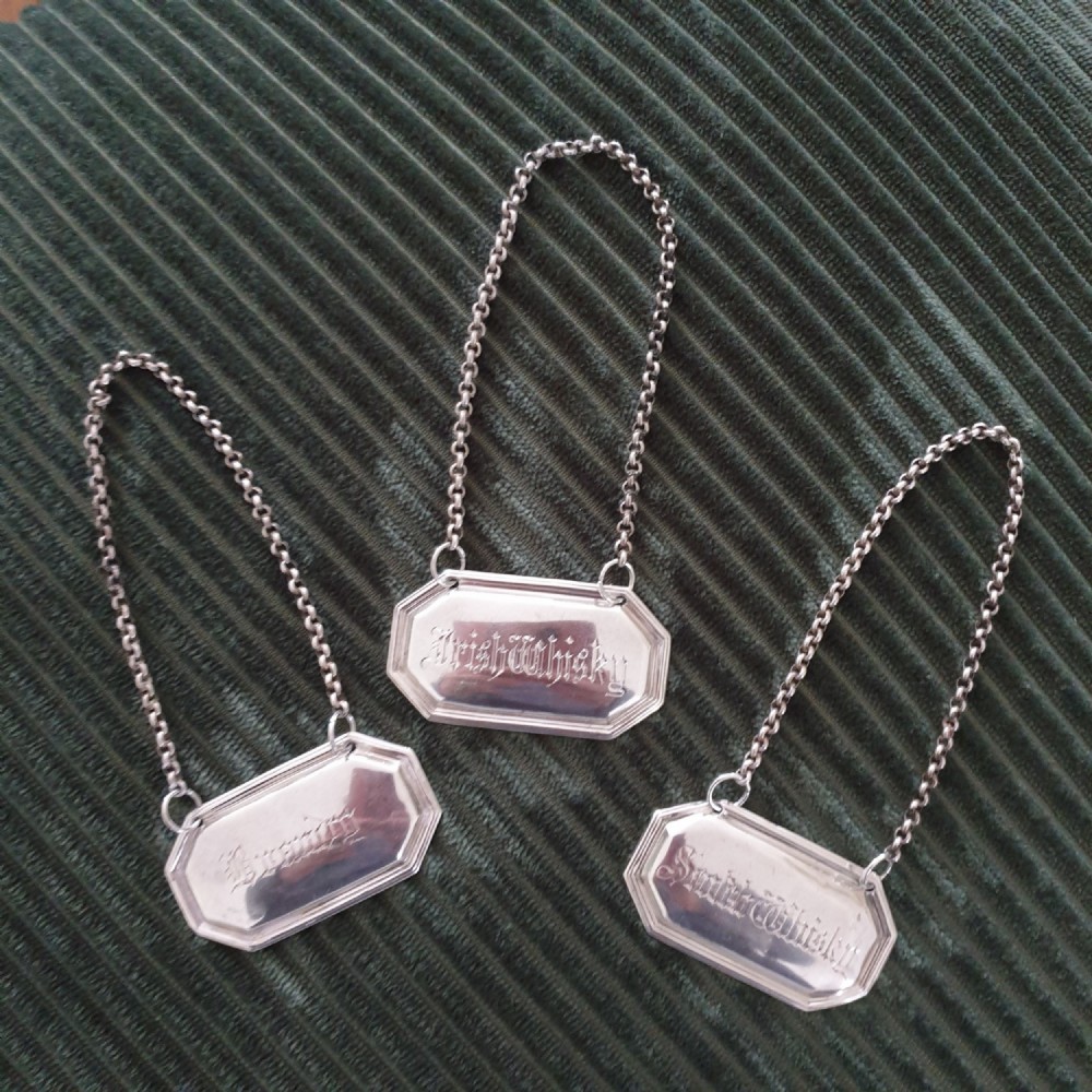 A set of three octagonal silver decanter labels with fine link chains, engraved with Brandy, Irish Whisky, and Scotch Whisky in Gothic script.
