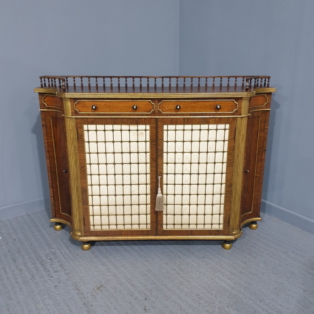 A Regency rosewood side cabinet featuring a spindle gallery rail, gilt brass trim, upper drawers, and fabric panel doors with brass lattice.