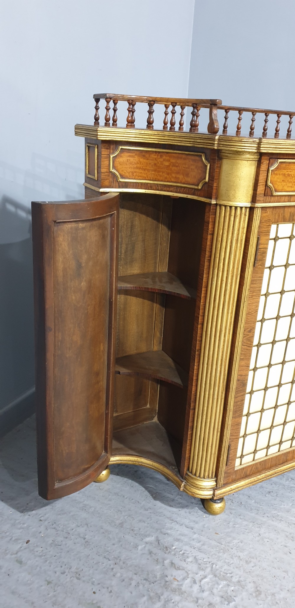 A Regency rosewood side cabinet featuring a spindle gallery rail, gilt brass trim, upper drawers, and fabric panel doors with brass lattice. View V