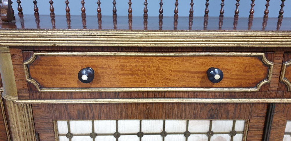 A Regency rosewood side cabinet featuring a spindle gallery rail, gilt brass trim, upper drawers, and fabric panel doors with brass lattice. View IX