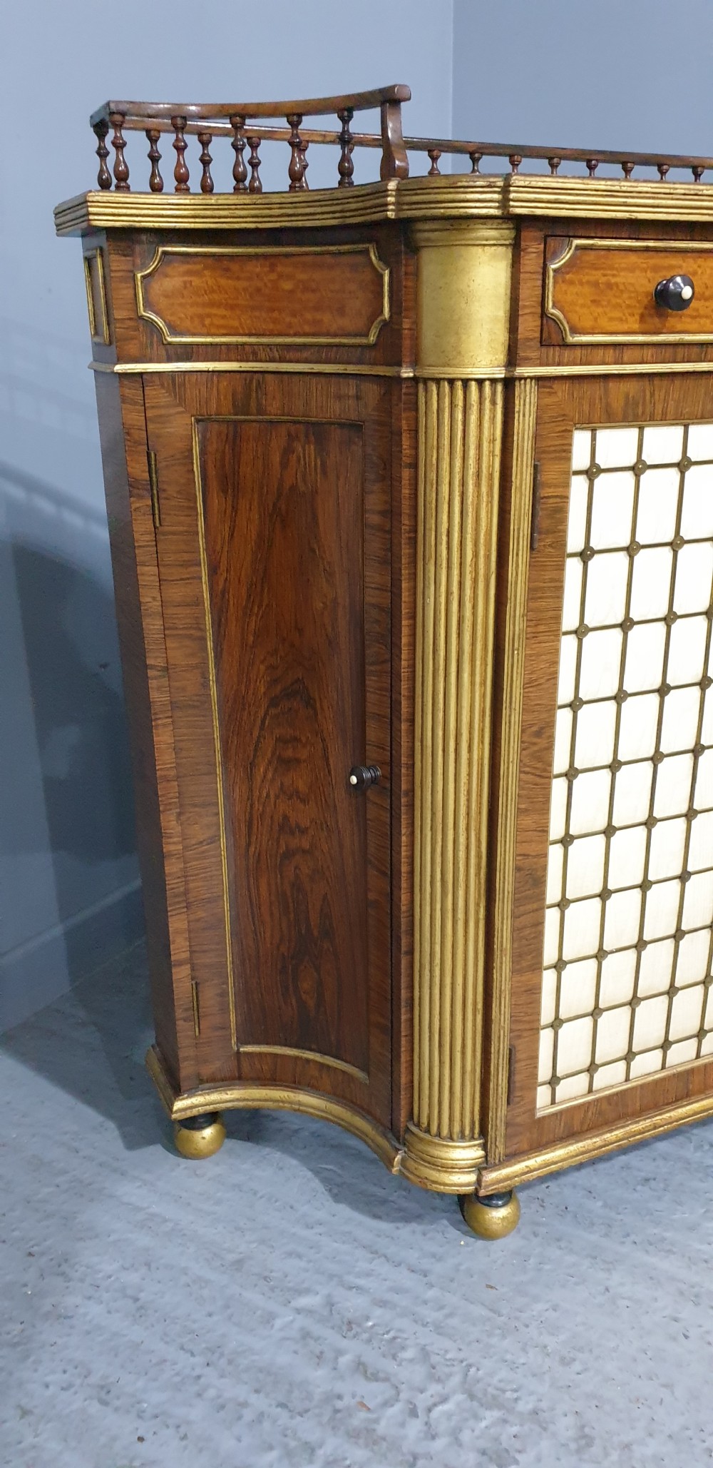 A Regency rosewood side cabinet featuring a spindle gallery rail, gilt brass trim, upper drawers, and fabric panel doors with brass lattice. View XIV