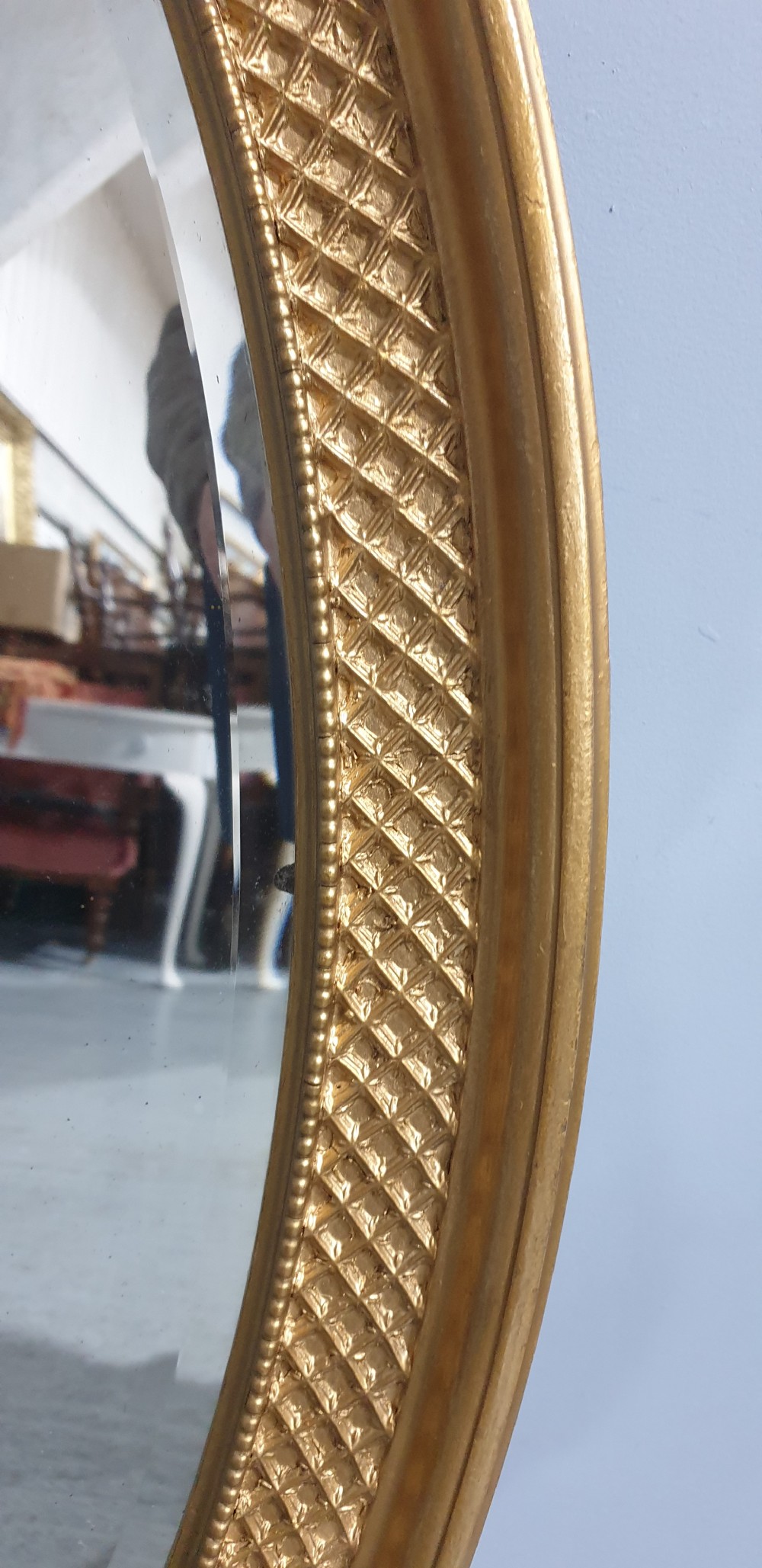 An oval Victorian wall mirror featuring a gilt gold frame with a textured lattice pattern surrounding the reflective glass mirror plate. View III