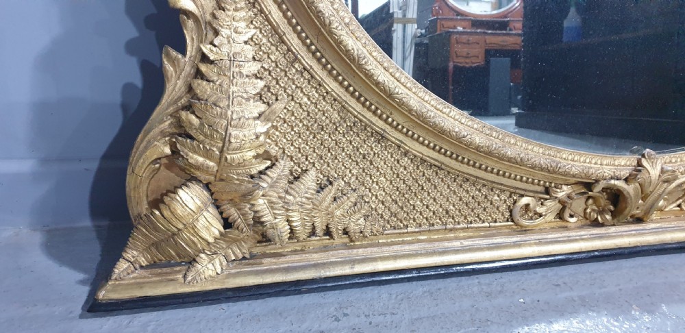 A large High Victorian arched overmantel mirror featuring a gold gilded frame decorated with carved fern leaf motifs, floral cresting, and geometric spandrel detailing. View VI