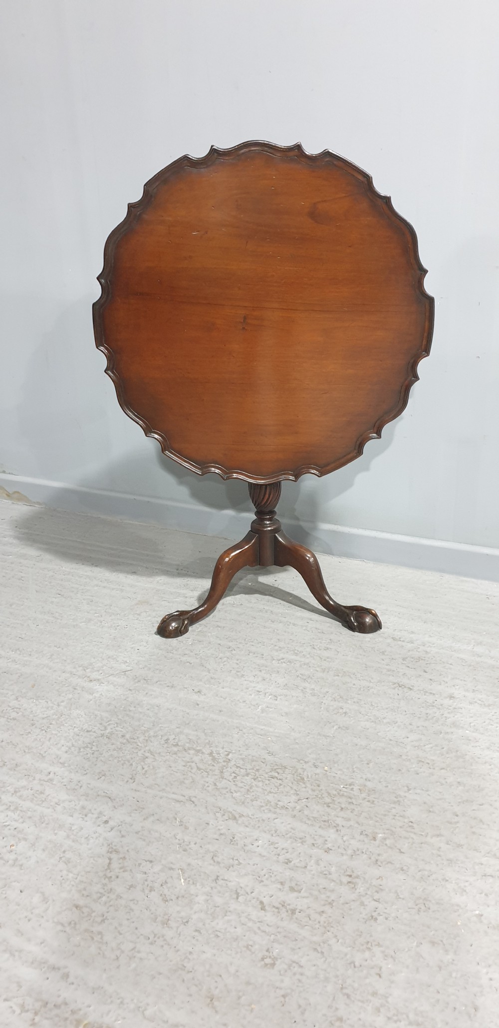 A Georgian style mahogany tilt-top table shown with the scalloped piecrust top folded vertically on a turned pedestal column with three carved legs.