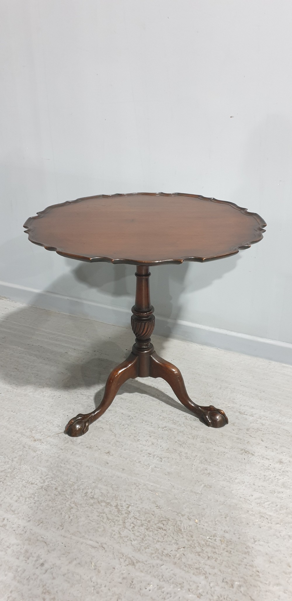 A Georgian style mahogany tilt-top table shown with the scalloped piecrust top folded vertically on a turned pedestal column with three carved legs. View II