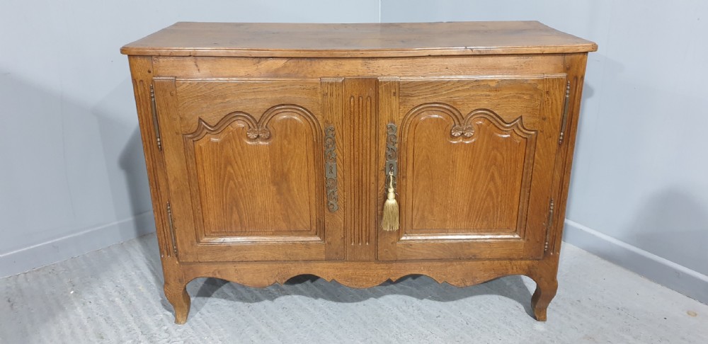 A French provincial oak dresser base featuring two carved arched cupboard doors, ornate metal keyhole hardware with a tassel, and short cabriole legs. View II