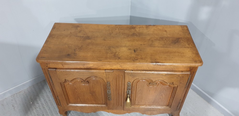 A French provincial oak dresser base featuring two carved arched cupboard doors, ornate metal keyhole hardware with a tassel, and short cabriole legs. View VII