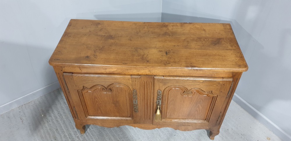 A French provincial oak dresser base featuring two carved arched cupboard doors, ornate metal keyhole hardware with a tassel, and short cabriole legs. View VIII