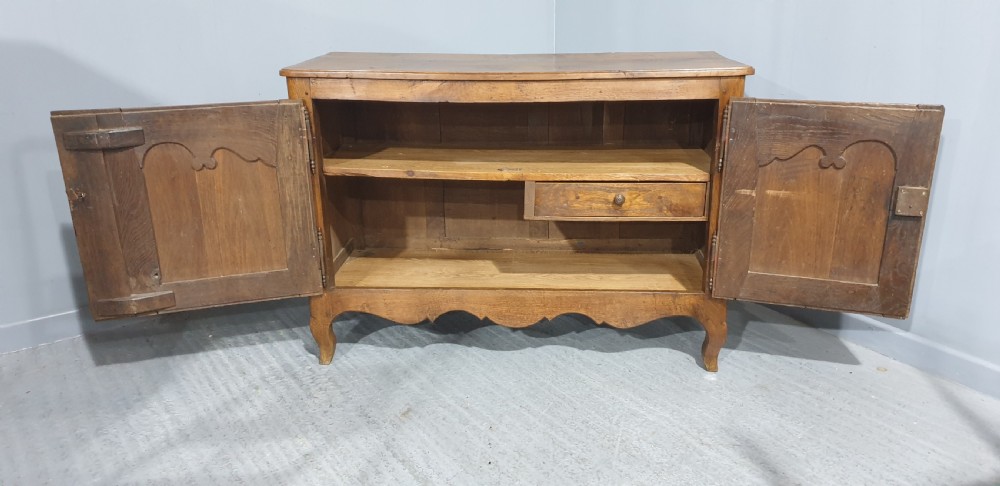 A French provincial oak dresser base featuring two carved arched cupboard doors, ornate metal keyhole hardware with a tassel, and short cabriole legs. View X