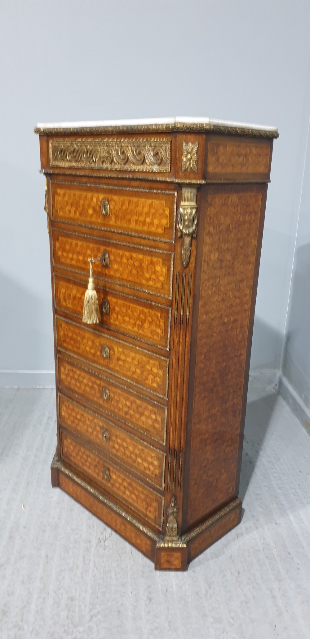 A tall French seven-drawer semainier chest with geometric wood parquetry, gold-toned ornamental mounts, and a white marble top. View V