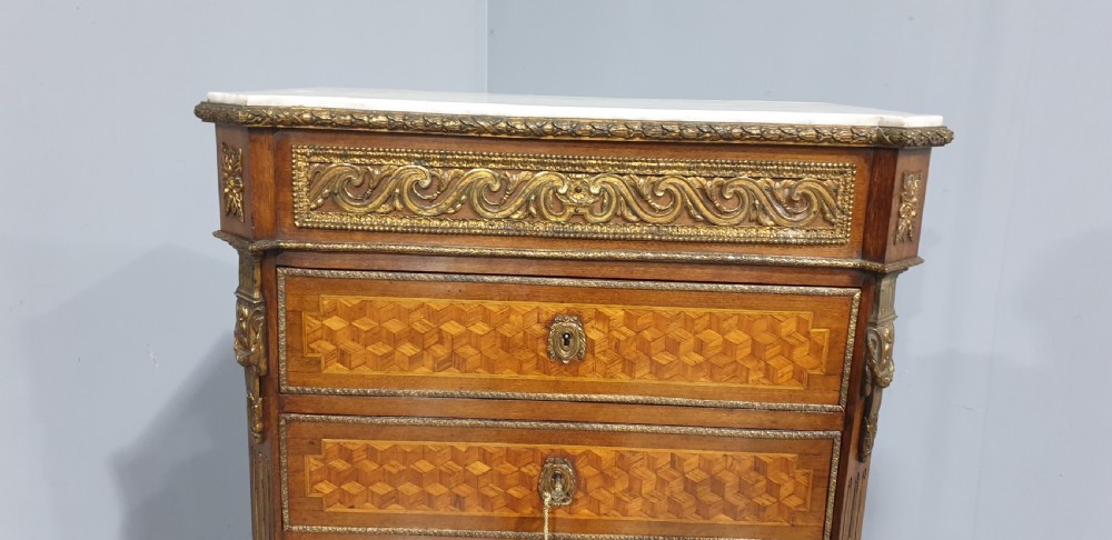 A tall French seven-drawer semainier chest with geometric wood parquetry, gold-toned ornamental mounts, and a white marble top. View VI