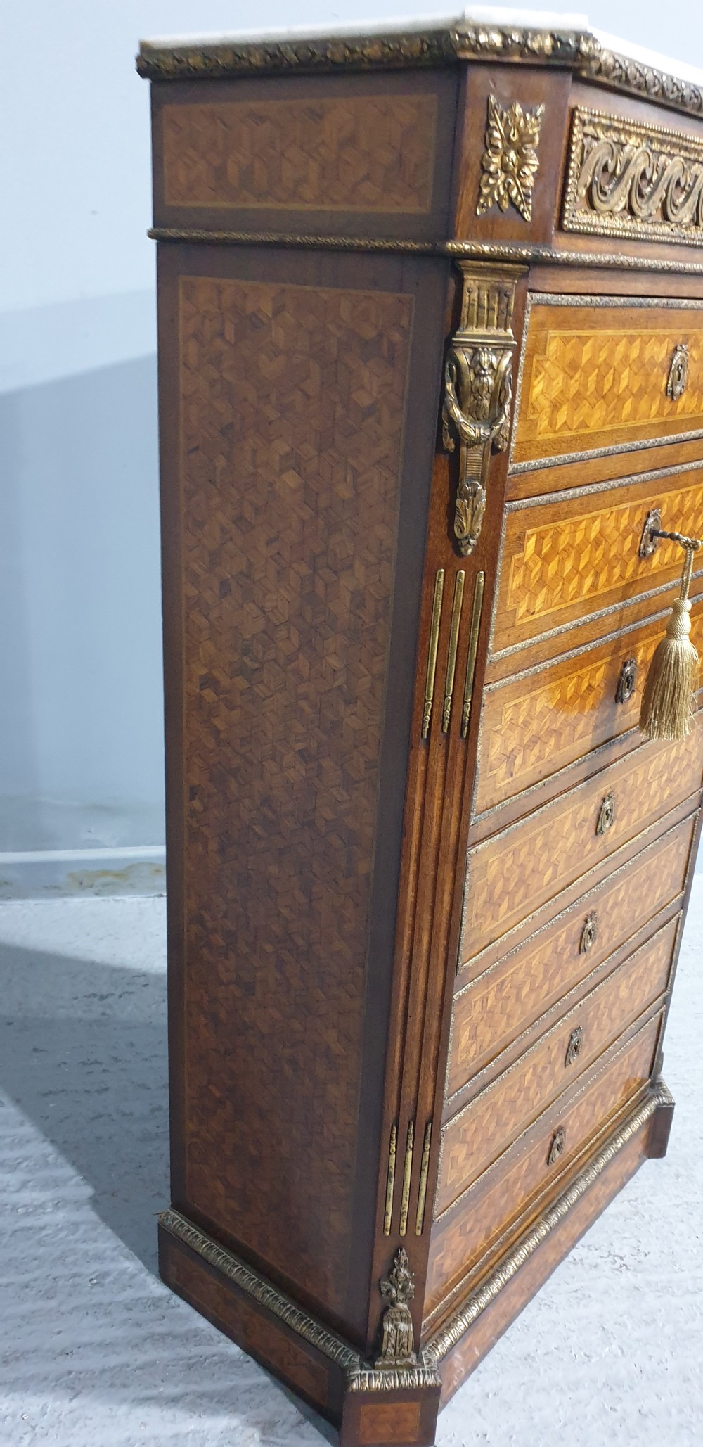 A tall French seven-drawer semainier chest with geometric wood parquetry, gold-toned ornamental mounts, and a white marble top. View VII