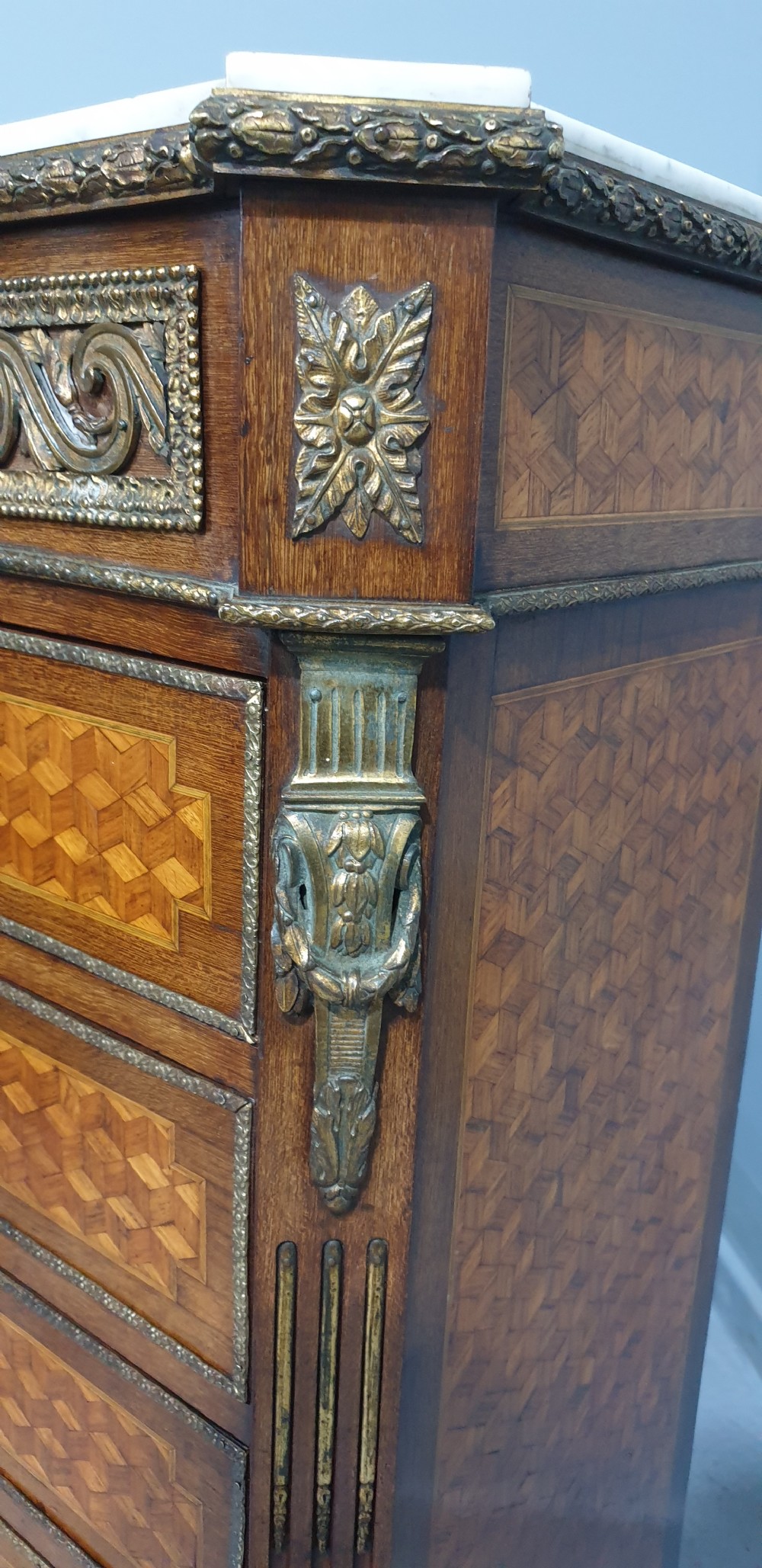 A tall French seven-drawer semainier chest with geometric wood parquetry, gold-toned ornamental mounts, and a white marble top. View IX