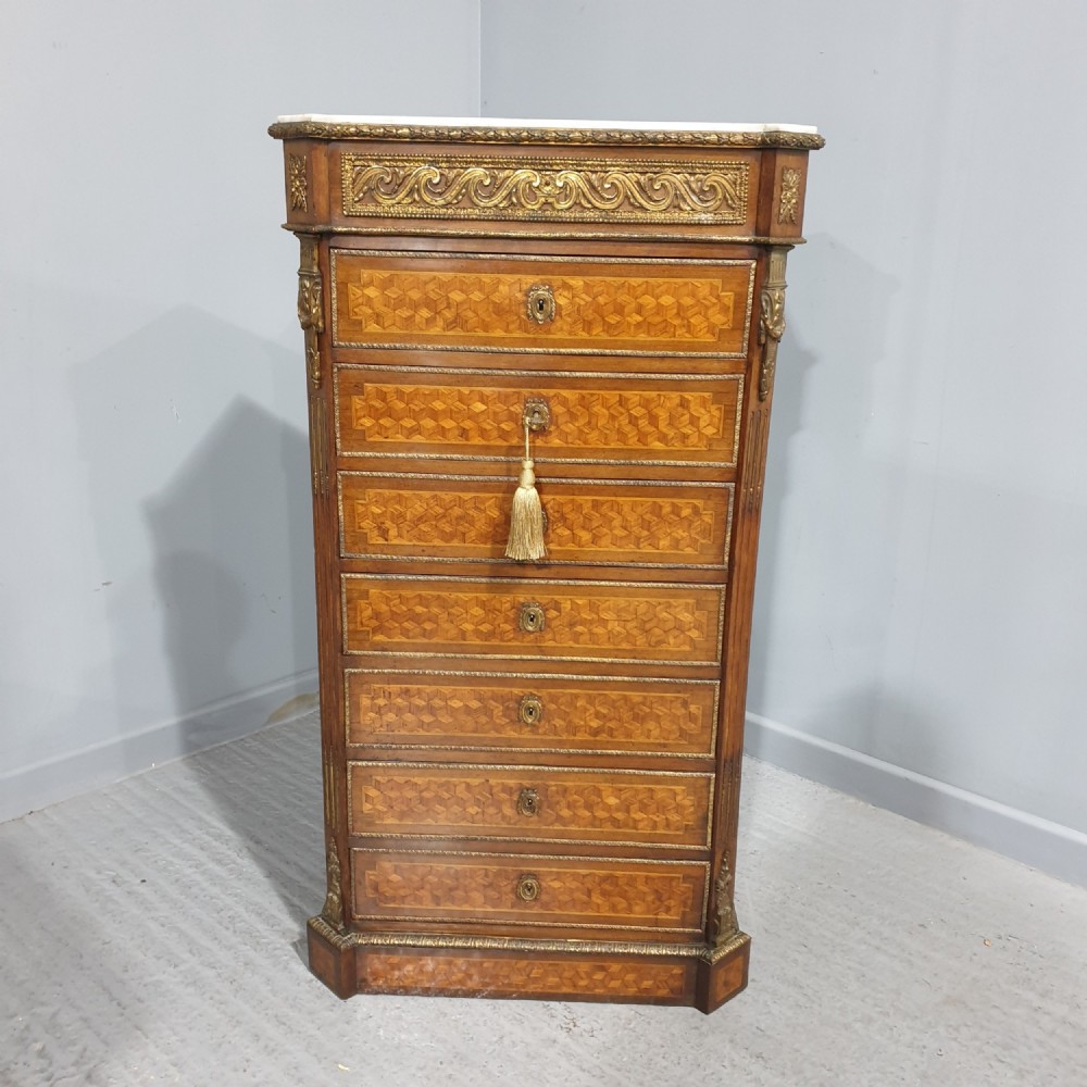 A tall French seven-drawer semainier chest with geometric wood parquetry, gold-toned ornamental mounts, and a white marble top.