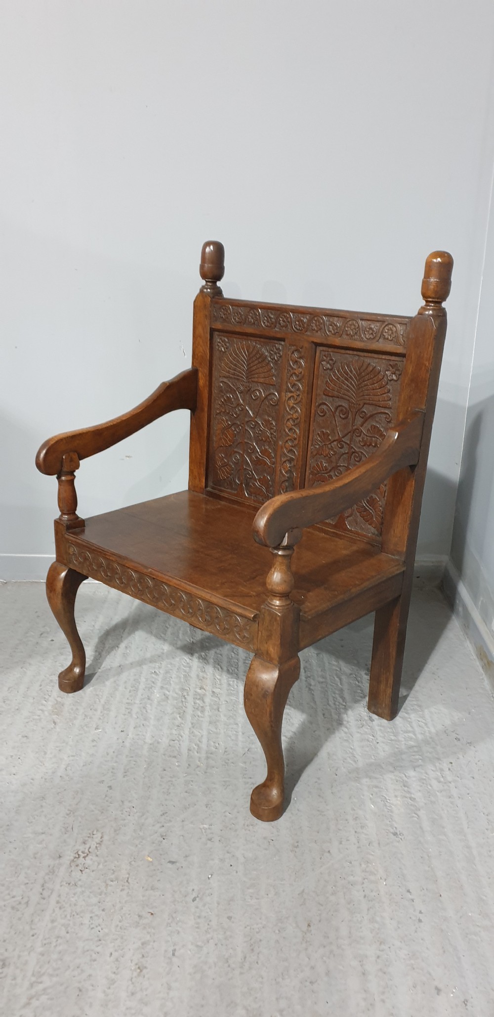 A small carved oak settle featuring a floral relief panelled back, turned finials, shaped armrests, and cabriole front legs.