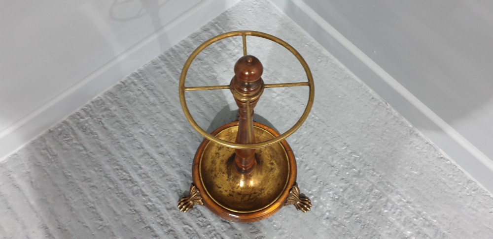 A Victorian turned mahogany stick stand featuring a circular top brass divider ring, a central turned stem, and a round brass drip tray base elevated by brass claw feet. View V