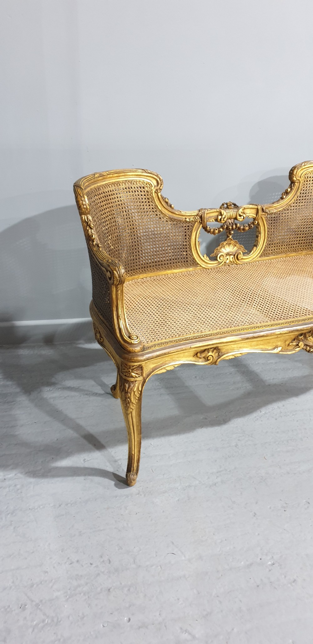 A Louis XV style French giltwood two-seater window bench featuring carved Rococo cartouches, cabriole legs, and natural woven cane panels. View V
