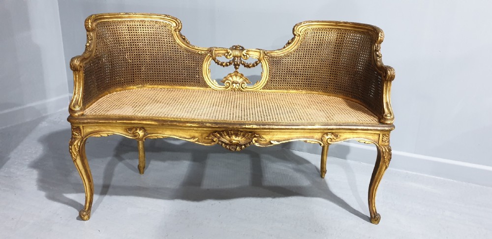 A Louis XV style French giltwood two-seater window bench featuring carved Rococo cartouches, cabriole legs, and natural woven cane panels. View X