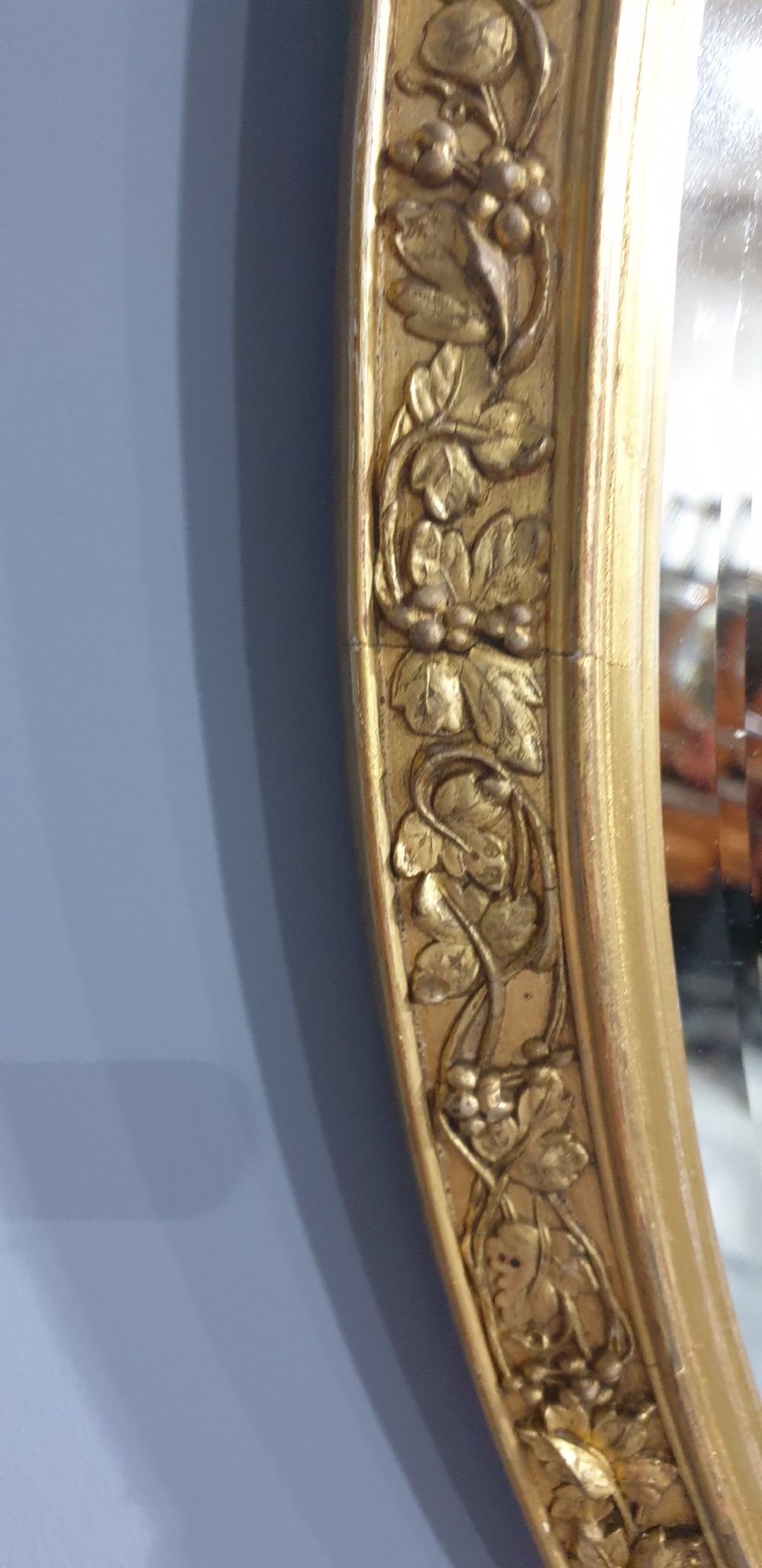 An oval wall mirror enclosed in a gilded frame featuring moulded floral relief ornamentation. View III