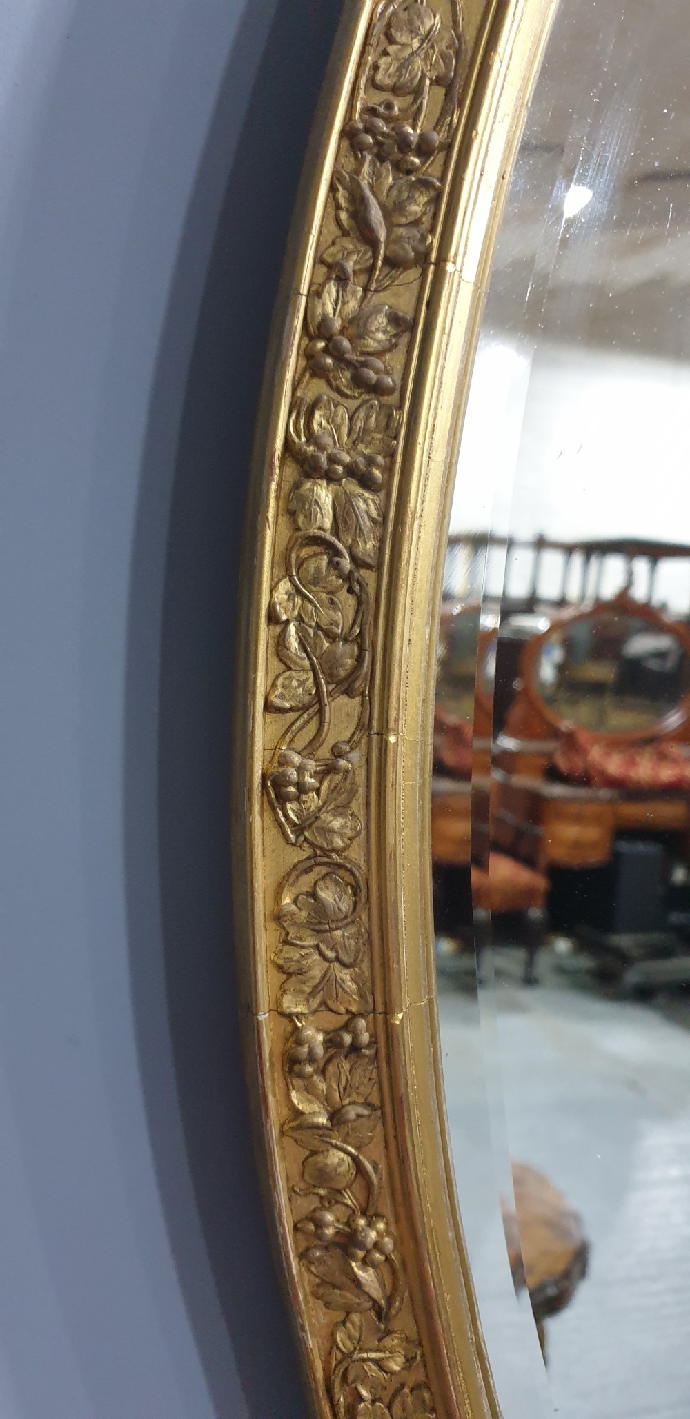 An oval wall mirror enclosed in a gilded frame featuring moulded floral relief ornamentation. View IV