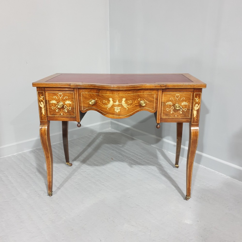 An antique rosewood writing desk featuring floral marquetry inlays, a red leather inset top, three drawers, and slender cabriole legs on brass castors.