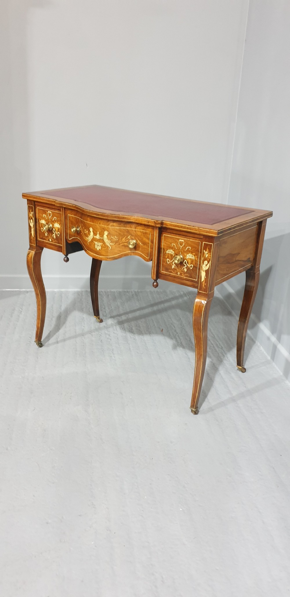 An antique rosewood writing desk featuring floral marquetry inlays, a red leather inset top, three drawers, and slender cabriole legs on brass castors. View II