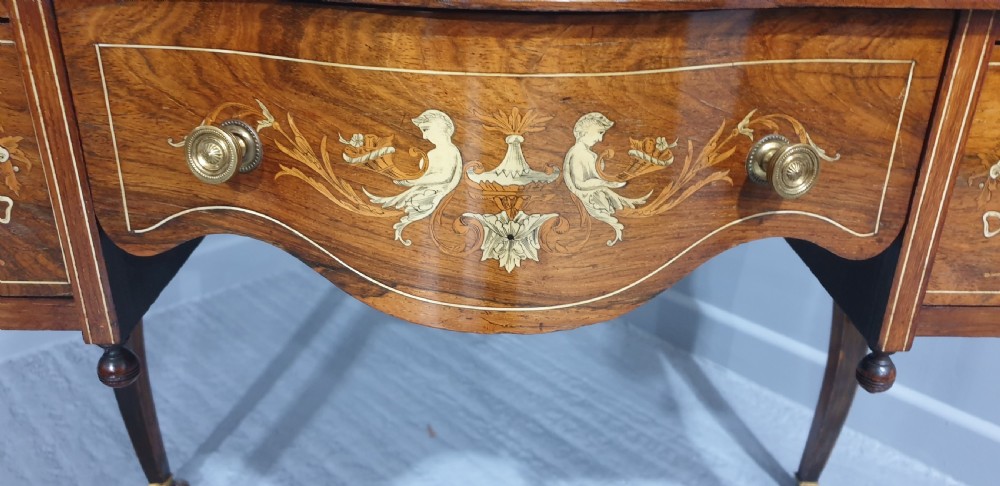 An antique rosewood writing desk featuring floral marquetry inlays, a red leather inset top, three drawers, and slender cabriole legs on brass castors. View V