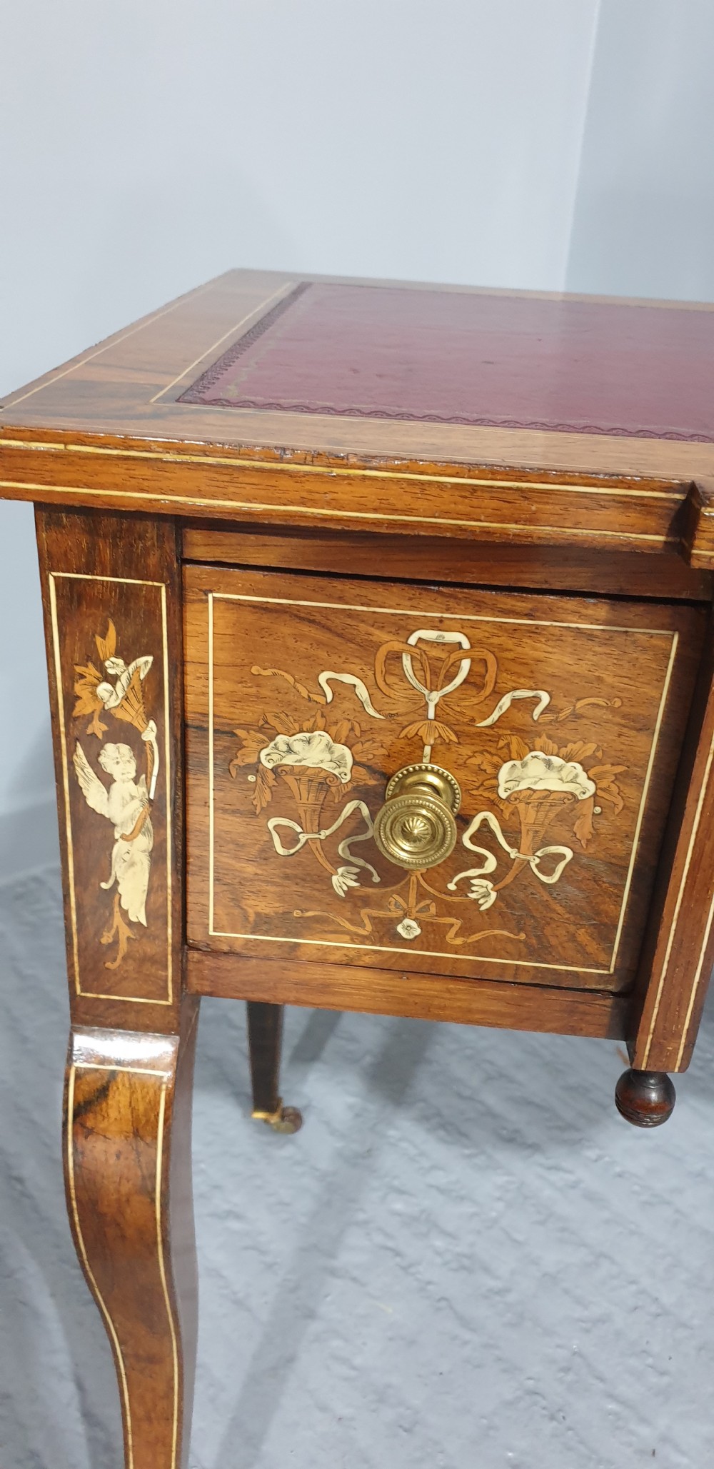 An antique rosewood writing desk featuring floral marquetry inlays, a red leather inset top, three drawers, and slender cabriole legs on brass castors. View VI