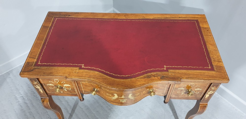 An antique rosewood writing desk featuring floral marquetry inlays, a red leather inset top, three drawers, and slender cabriole legs on brass castors. View VII