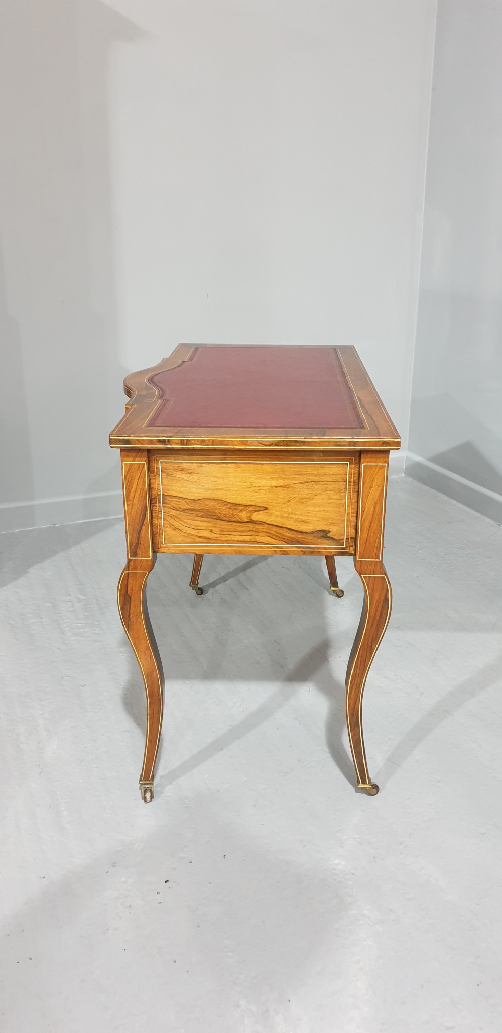 An antique rosewood writing desk featuring floral marquetry inlays, a red leather inset top, three drawers, and slender cabriole legs on brass castors. View VIII
