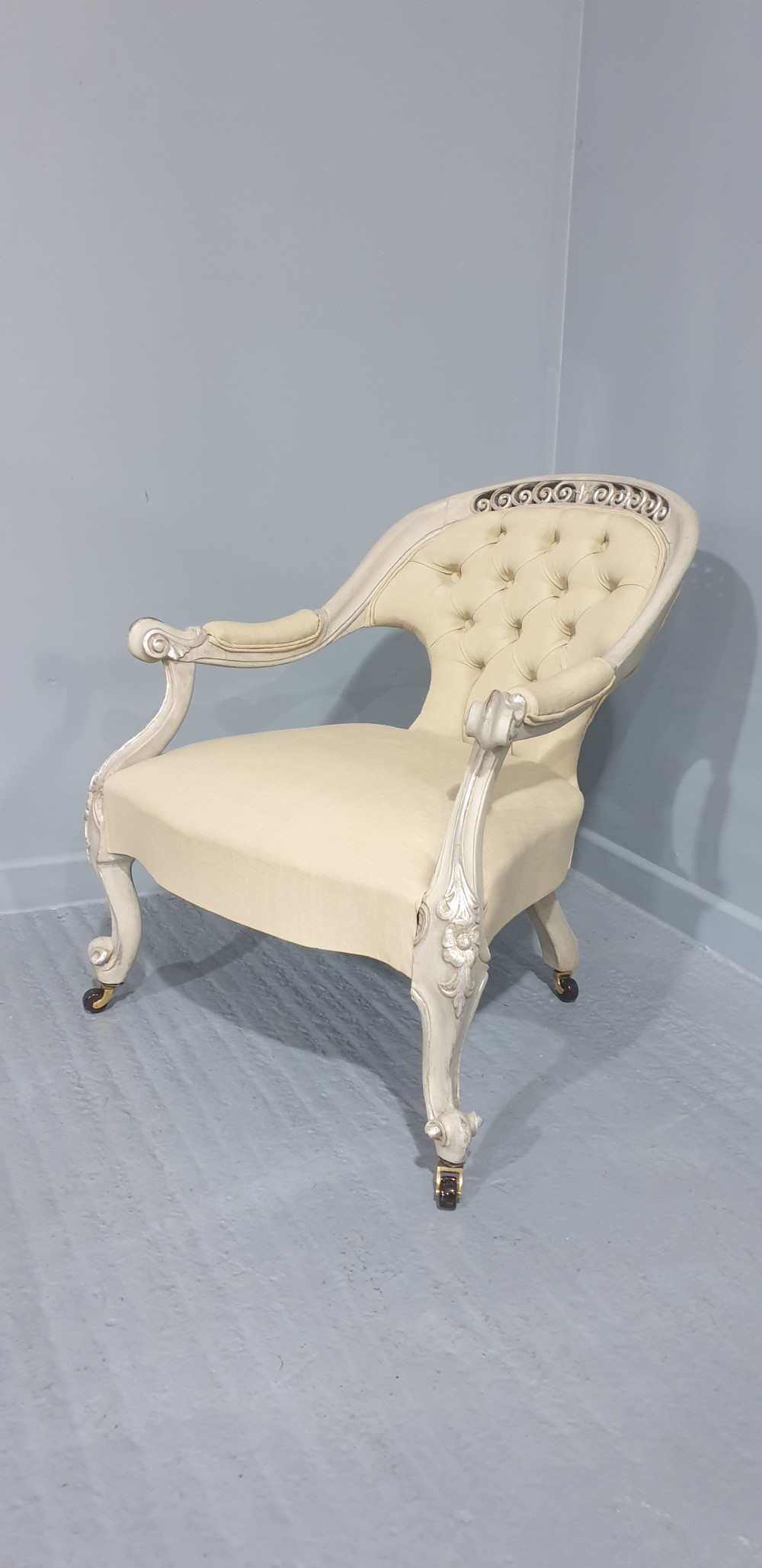 A Victorian low-seated armchair with a carved off-white frame, silver gilt highlights, and button-tufted cream fabric upholstery resting on front brass castors. View III