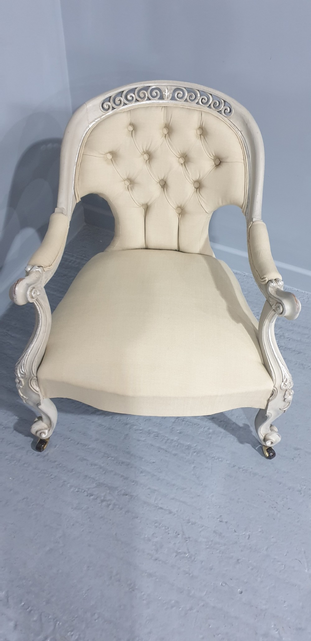 A Victorian low-seated armchair with a carved off-white frame, silver gilt highlights, and button-tufted cream fabric upholstery resting on front brass castors. View IV