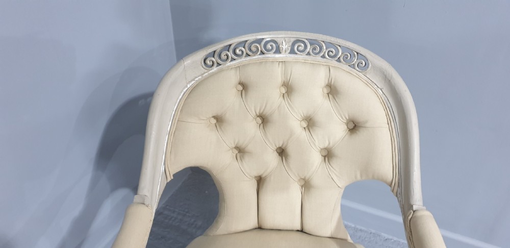 A Victorian low-seated armchair with a carved off-white frame, silver gilt highlights, and button-tufted cream fabric upholstery resting on front brass castors. View VI