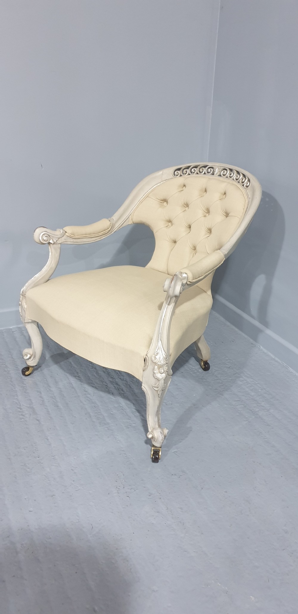 A Victorian low-seated armchair with a carved off-white frame, silver gilt highlights, and button-tufted cream fabric upholstery resting on front brass castors. View VII