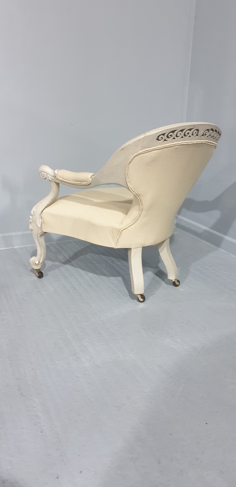 A Victorian low-seated armchair with a carved off-white frame, silver gilt highlights, and button-tufted cream fabric upholstery resting on front brass castors. View IX