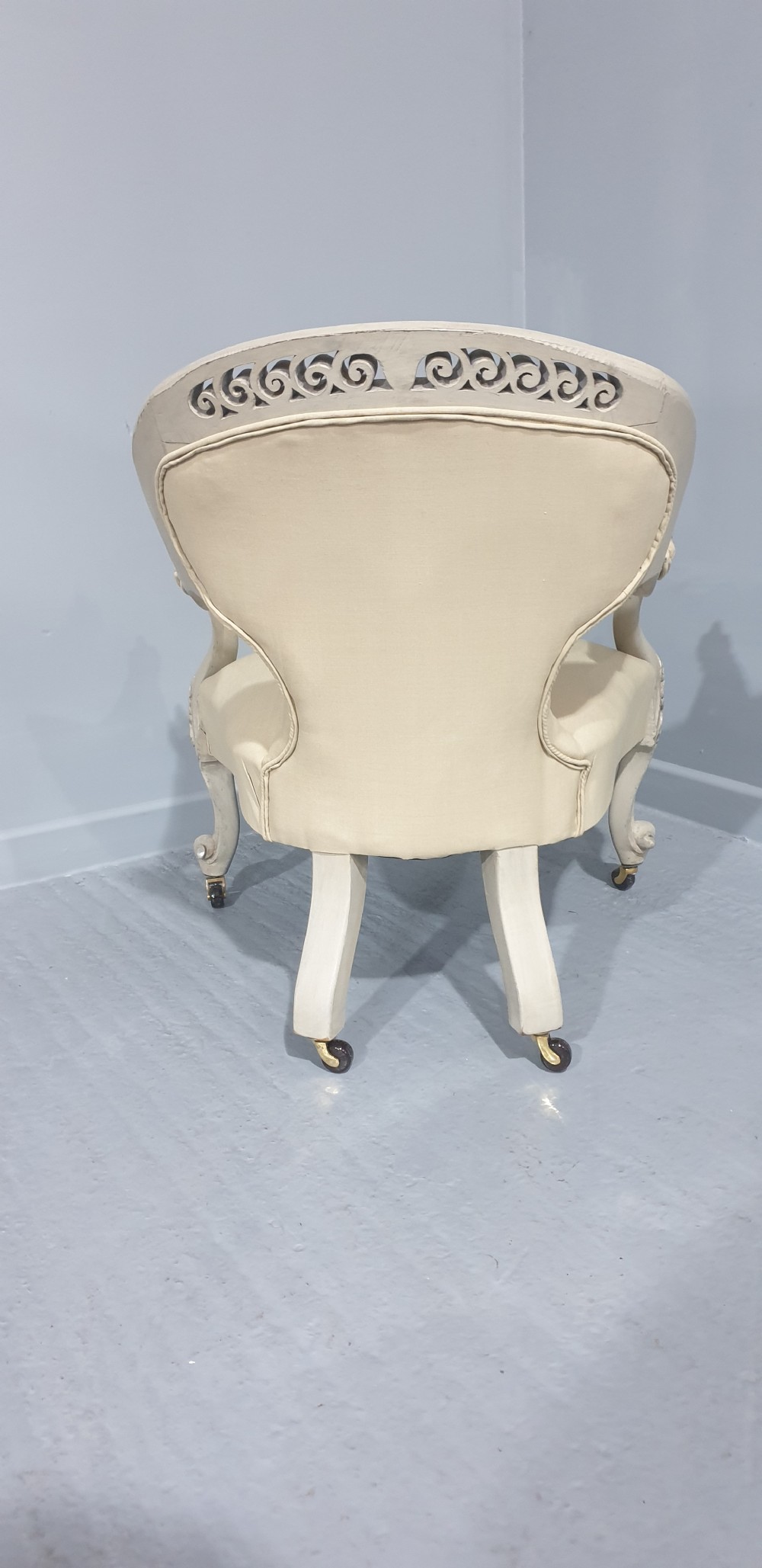 A Victorian low-seated armchair with a carved off-white frame, silver gilt highlights, and button-tufted cream fabric upholstery resting on front brass castors. View X