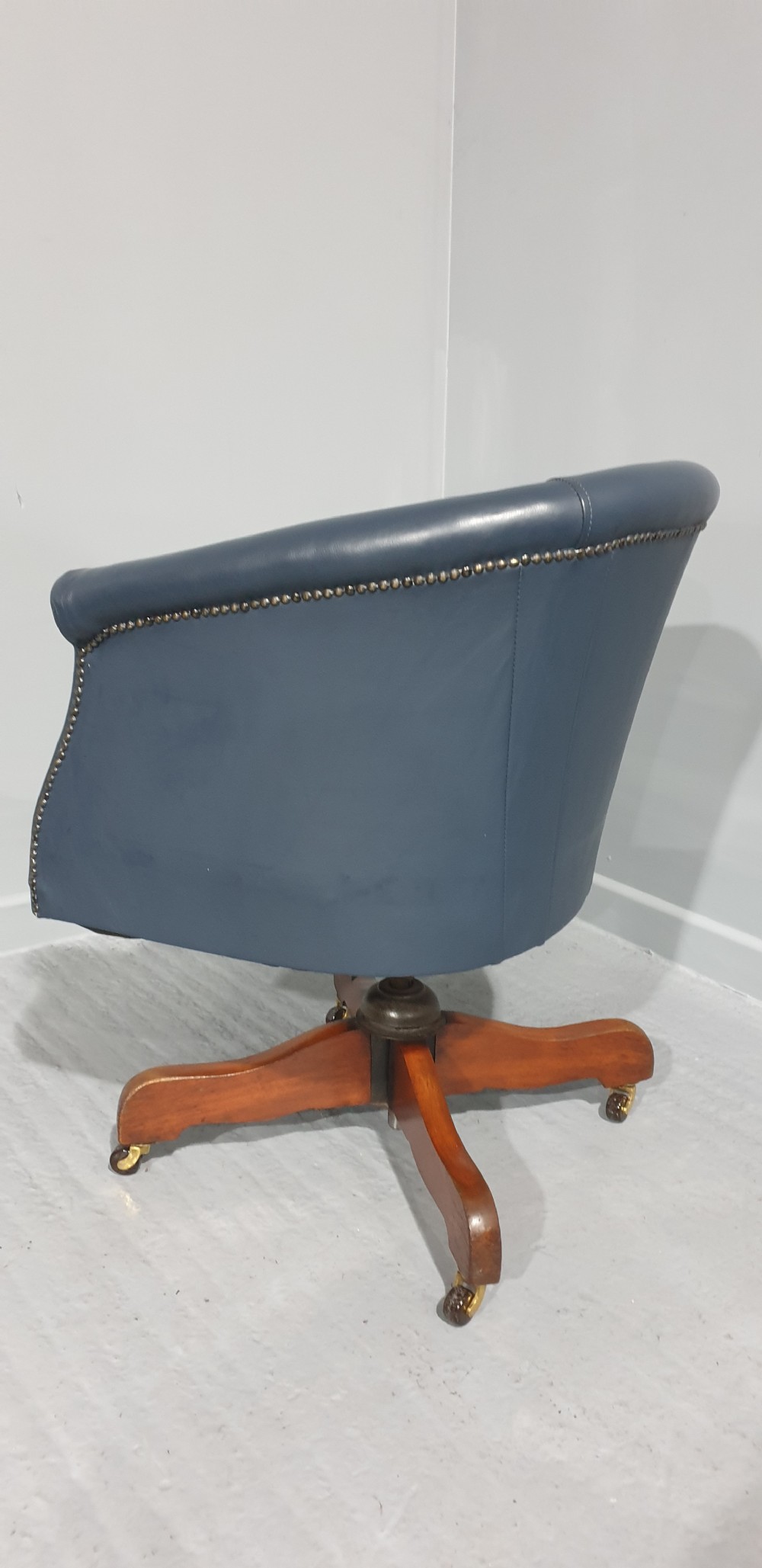 A blue leather tub desk chair with a curved backrest, brass studding detail, and a swivel wood quadruped base on castors. View V
