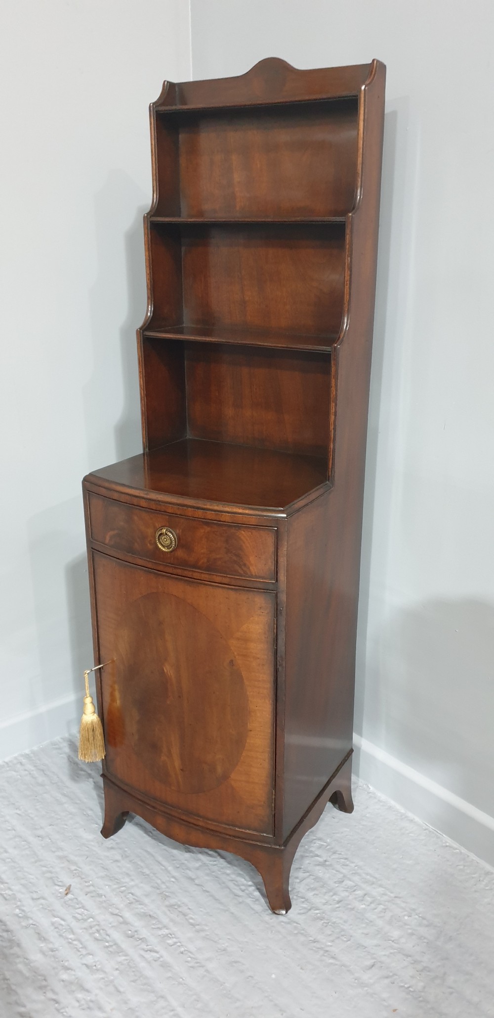 A tall, narrow mahogany cabinet featuring three open graduated shelves above a single drawer and a lower cupboard door with an oval flame mahogany inlay. View X