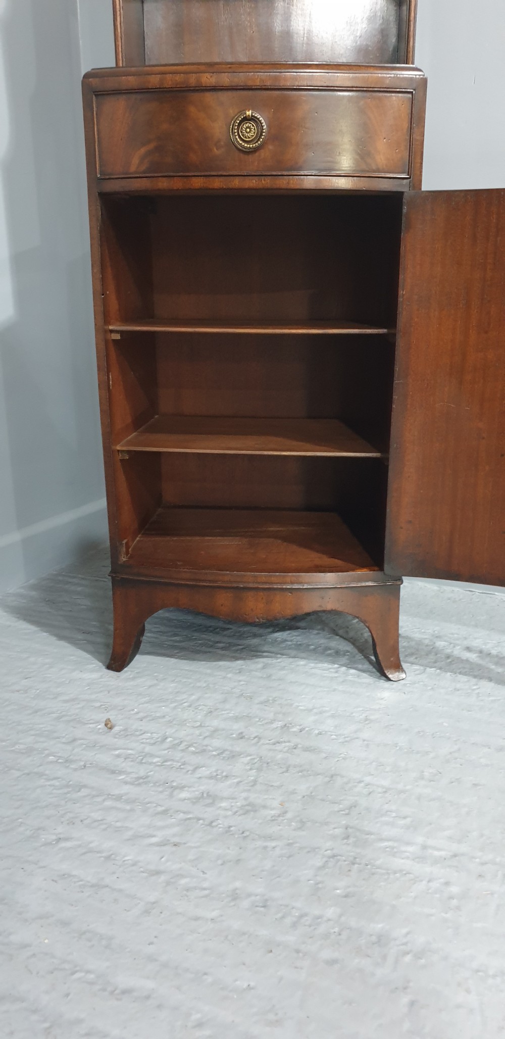 A tall, narrow mahogany cabinet featuring three open graduated shelves above a single drawer and a lower cupboard door with an oval flame mahogany inlay. View XI