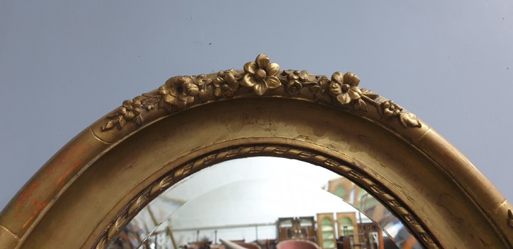 An oval Victorian wall mirror featuring a giltwood frame decorated with a floral motif outer border and a inner rope twist moulding. View V