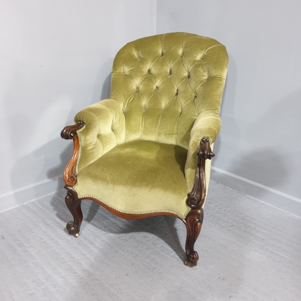 A Victorian library armchair with deep button-tufted olive green velvet upholstery, a carved mahogany frame, and cabriole legs on brass castors.