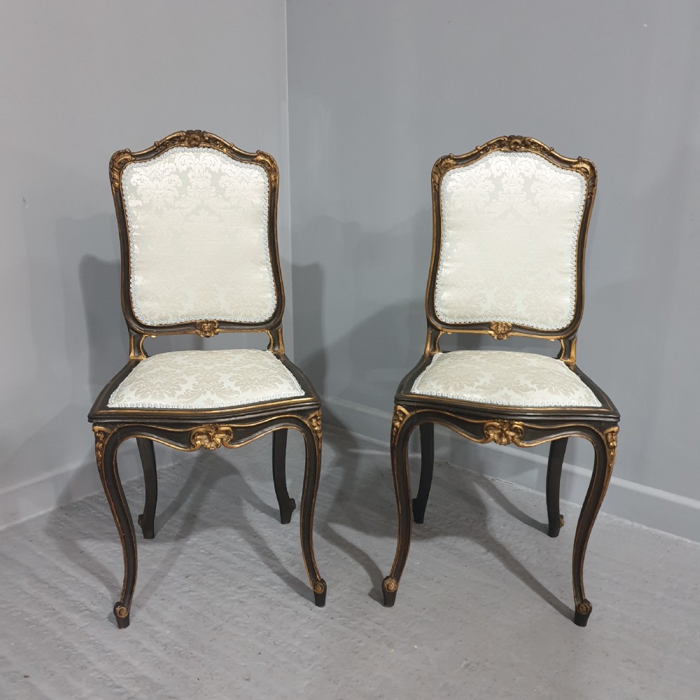 A pair of French Rococo style side chairs with dark ebonised and giltwood frames, carved cabriole legs, and cream damask upholstery.