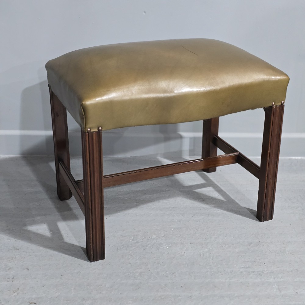 A Georgian mahogany stool featuring fluted square legs, an H-stretcher, and an olive green leather upholstered seat with brass stud details.