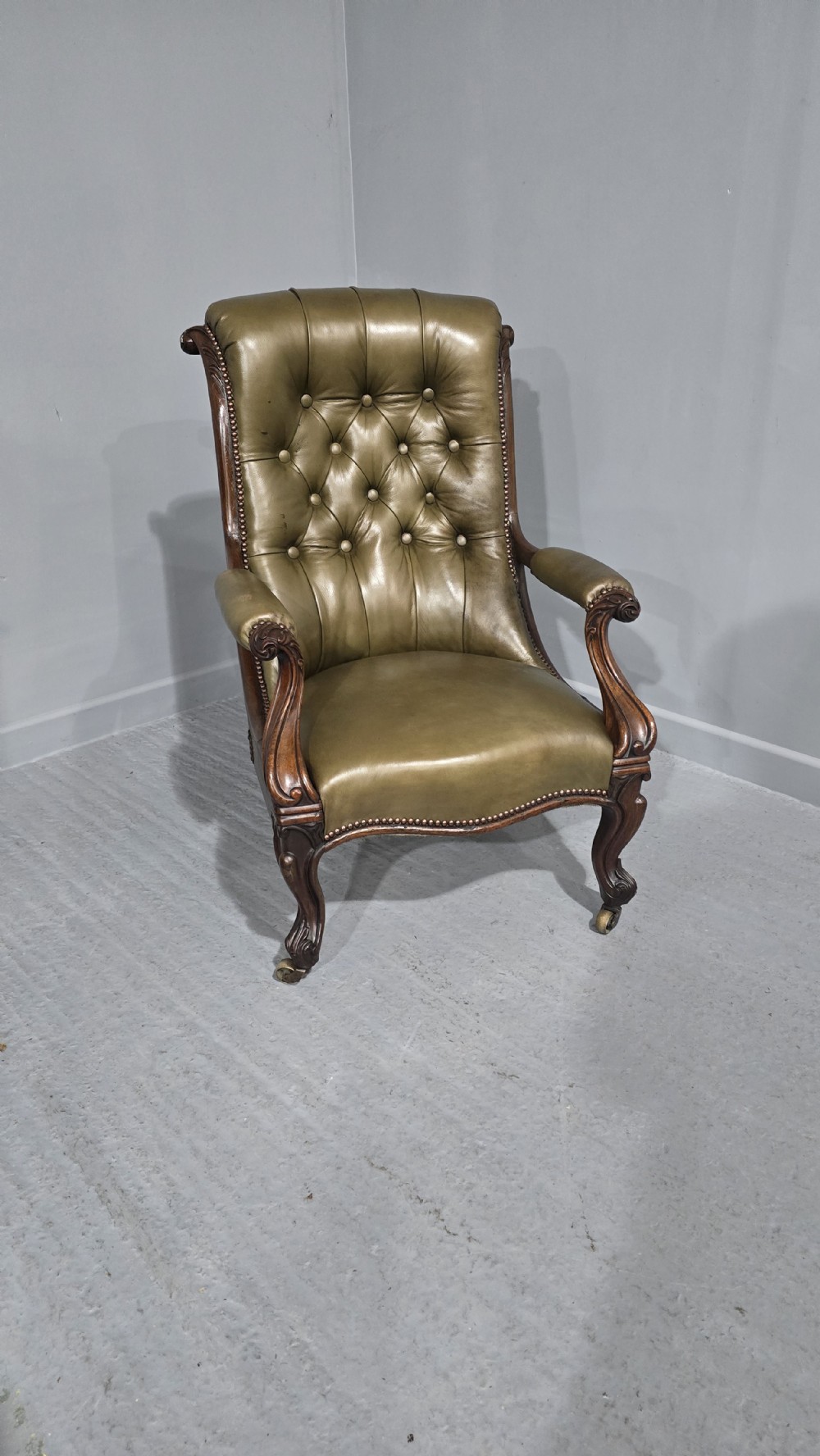 An antique Victorian green leather library chair with deep-buttoned upholstery, carved mahogany frame, scrolled arms, and cabriole front legs on brass castors.