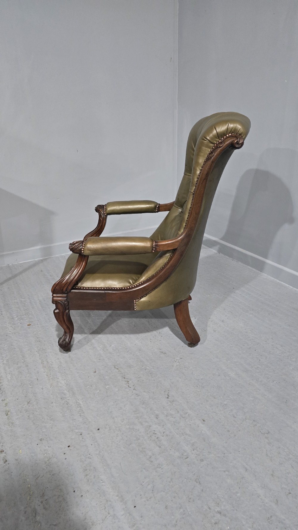 An antique Victorian green leather library chair with deep-buttoned upholstery, carved mahogany frame, scrolled arms, and cabriole front legs on brass castors. View II