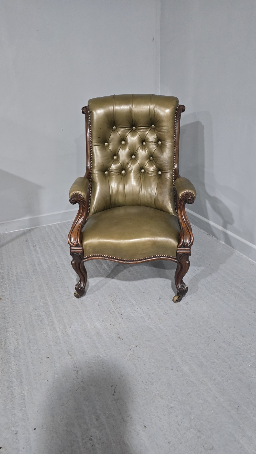 An antique Victorian green leather library chair with deep-buttoned upholstery, carved mahogany frame, scrolled arms, and cabriole front legs on brass castors. View III