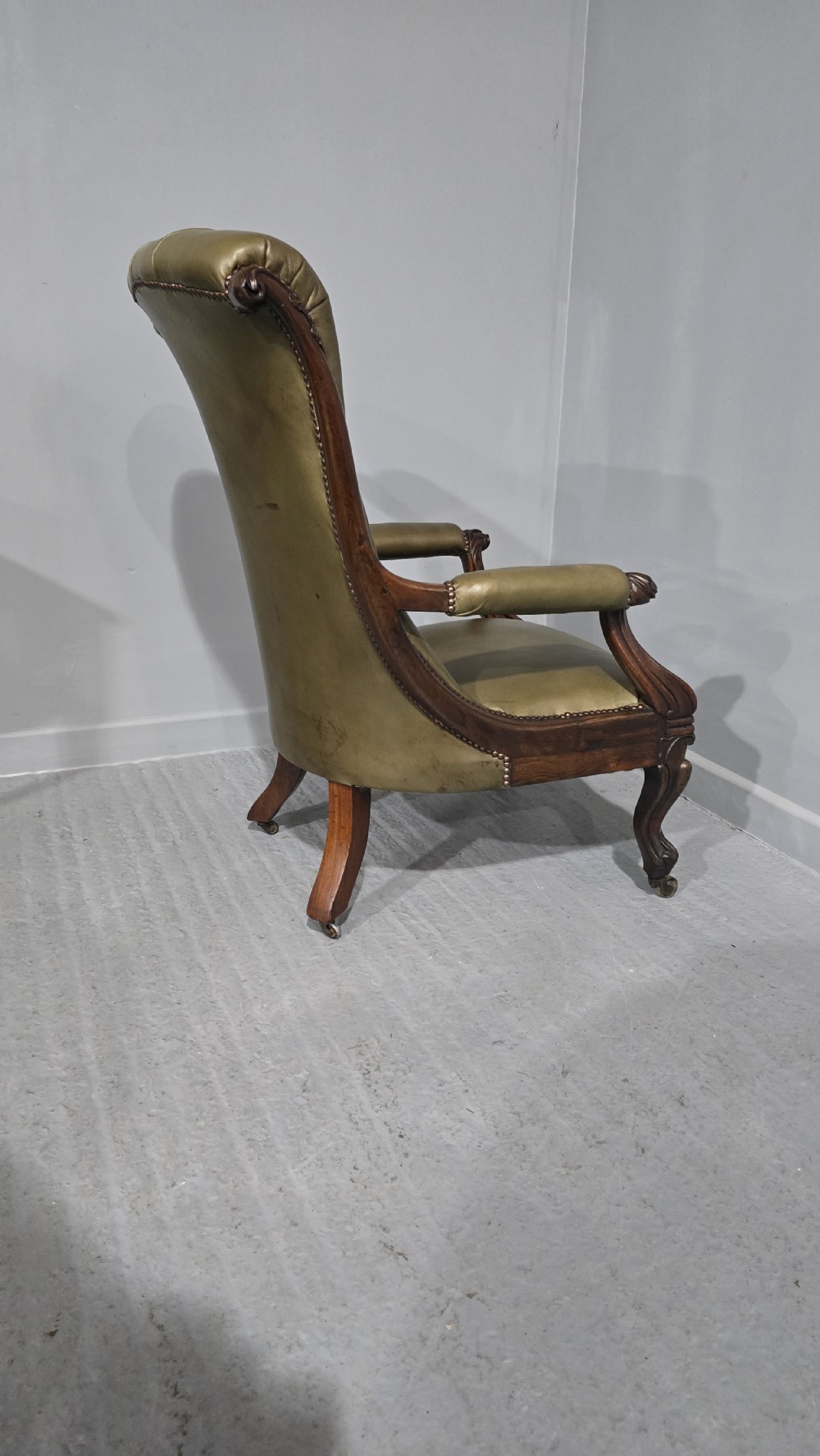 An antique Victorian green leather library chair with deep-buttoned upholstery, carved mahogany frame, scrolled arms, and cabriole front legs on brass castors. View VI