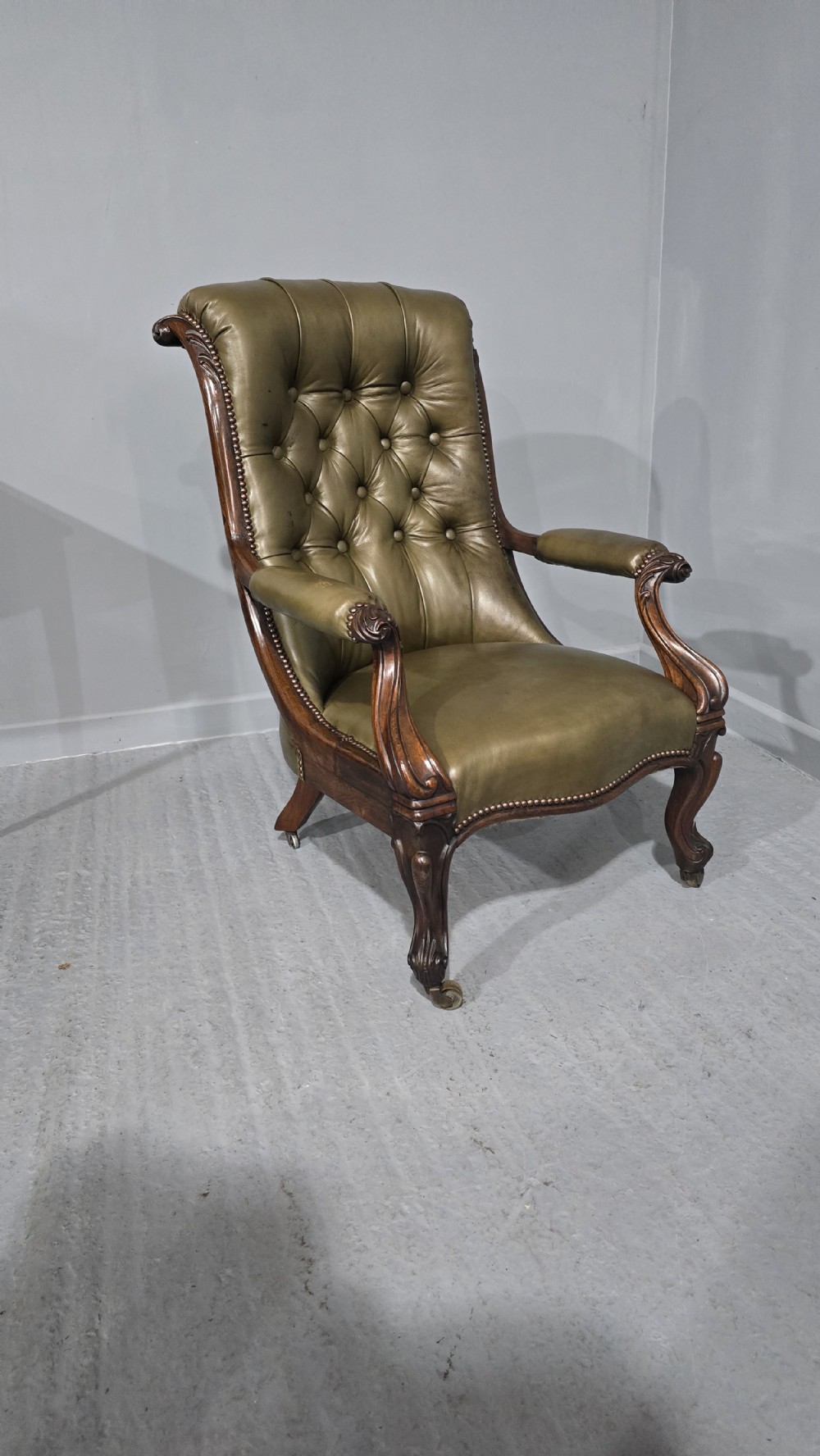 An antique Victorian green leather library chair with deep-buttoned upholstery, carved mahogany frame, scrolled arms, and cabriole front legs on brass castors. View VII